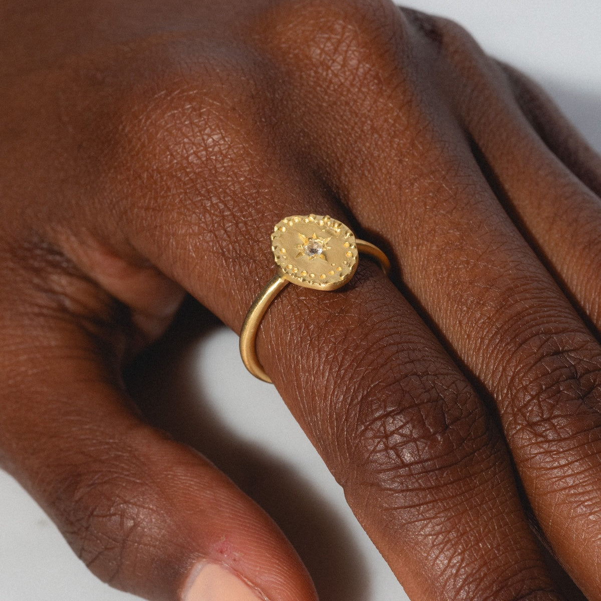 A hand with dark brown skin wears the Brahma Ring, a gold plated ring with an irregular disc and a white topaz at its center, on the middle finger.
