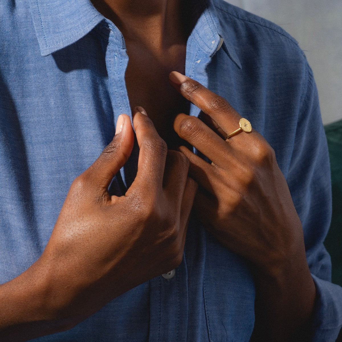 A person in a blue button-up shirt shows off the Brahma Ring, featuring a yellow gold plated finish and white topaz design, by holding their collar with one hand and touching their chest with the other.