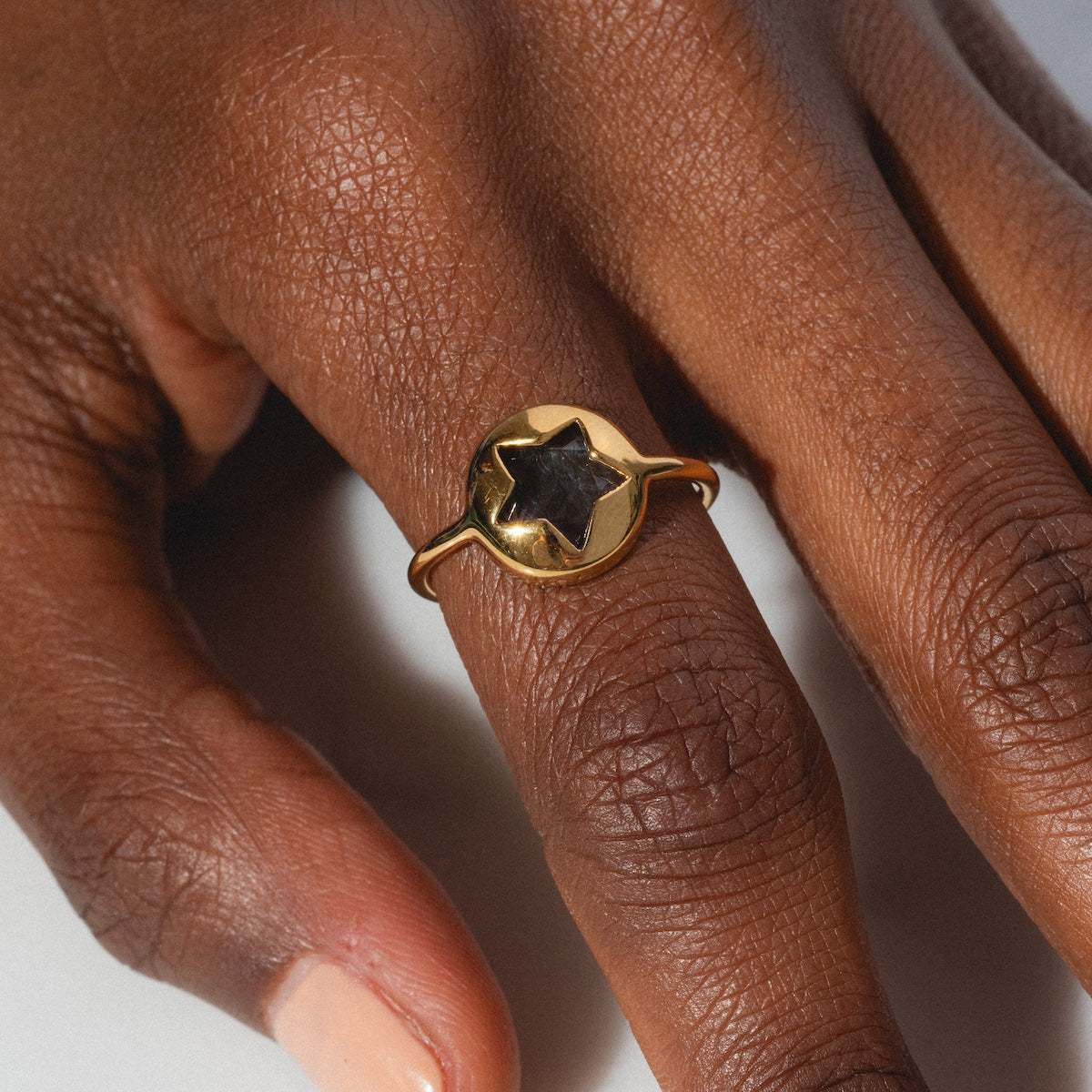 A close-up shows a medium-brown hand with short, natural nails wearing the Maomi Ring—yellow gold plated and set with a black star-shaped stone at the center.