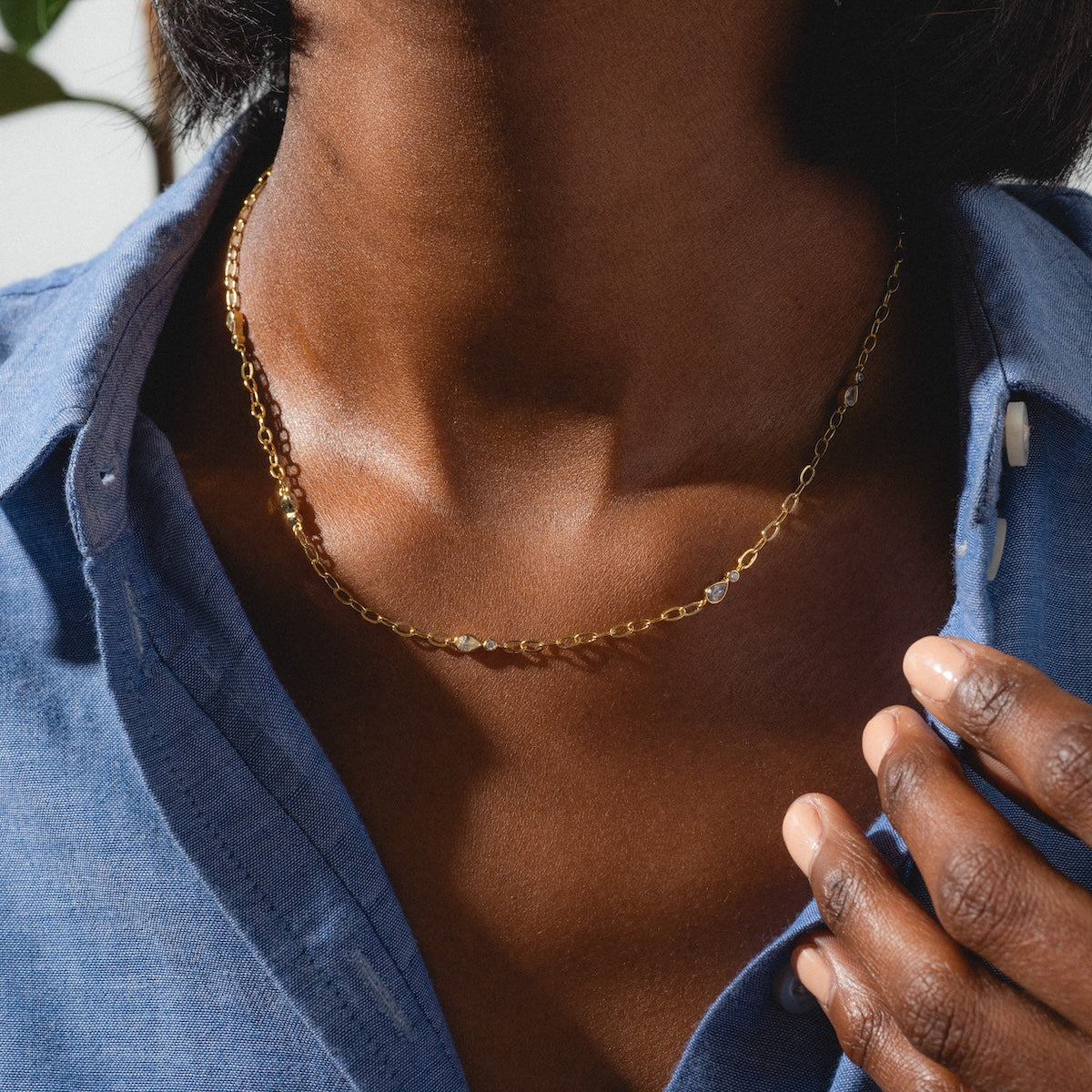 Wearing a blue shirt, a person touches their collarbone, showcasing the Perfect Paperclip Layering Necklace Sample in yellow gold plating. The lighting highlights both the necklace and the natural texture of their skin.