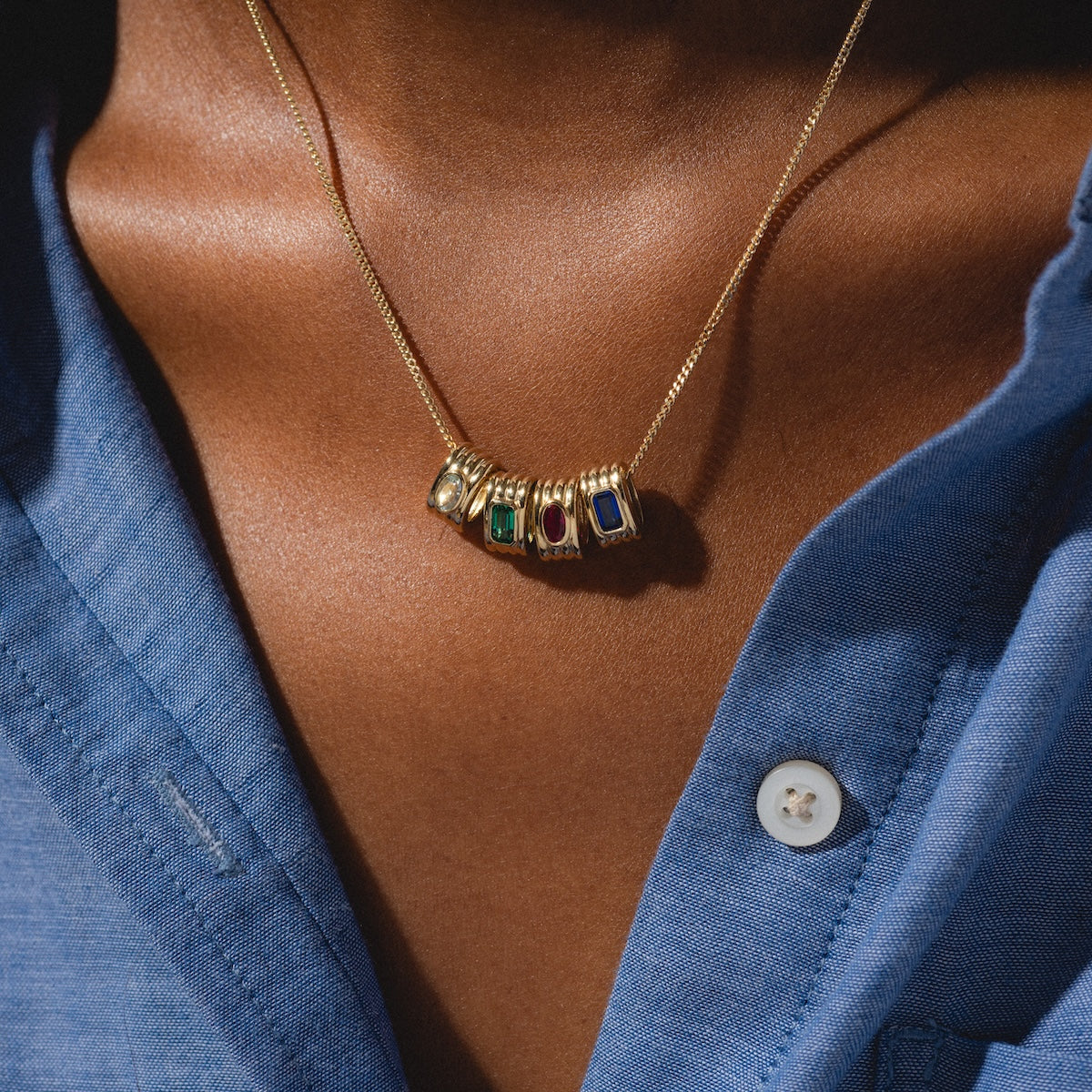 A close-up of a person wearing a Solid Gold Bold Birthstone Spacer Charm as a necklace pendant, paired with a blue button-up shirt.