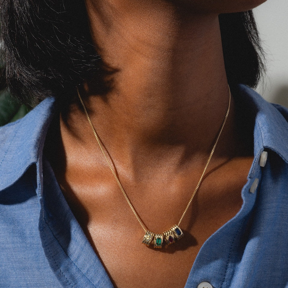 A woman in a blue button-up shirt wears the Solid Gold Bold Birthstone Spacer Charm necklace, featuring colorful beads, while posing against a blurred background with a green leaf partly visible.