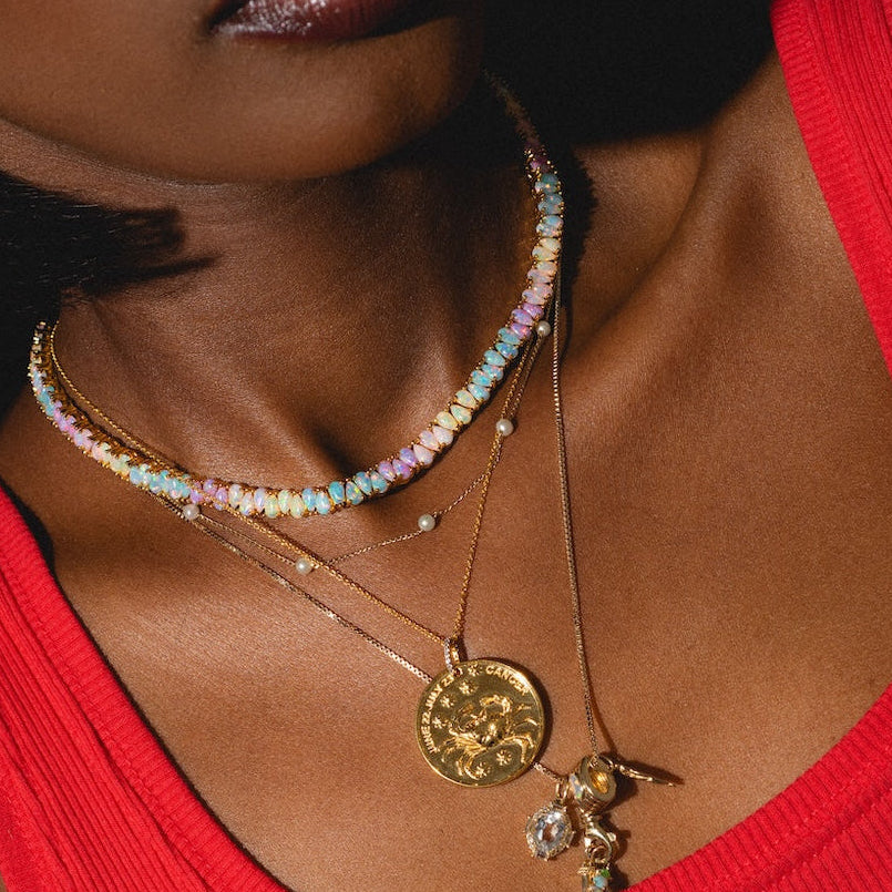 A close-up of a woman wearing a red tank top, layered gold necklaces, and the Solid Gold Bold Birthstone Spacer Charm, with warm lighting highlighting her neck and lips.