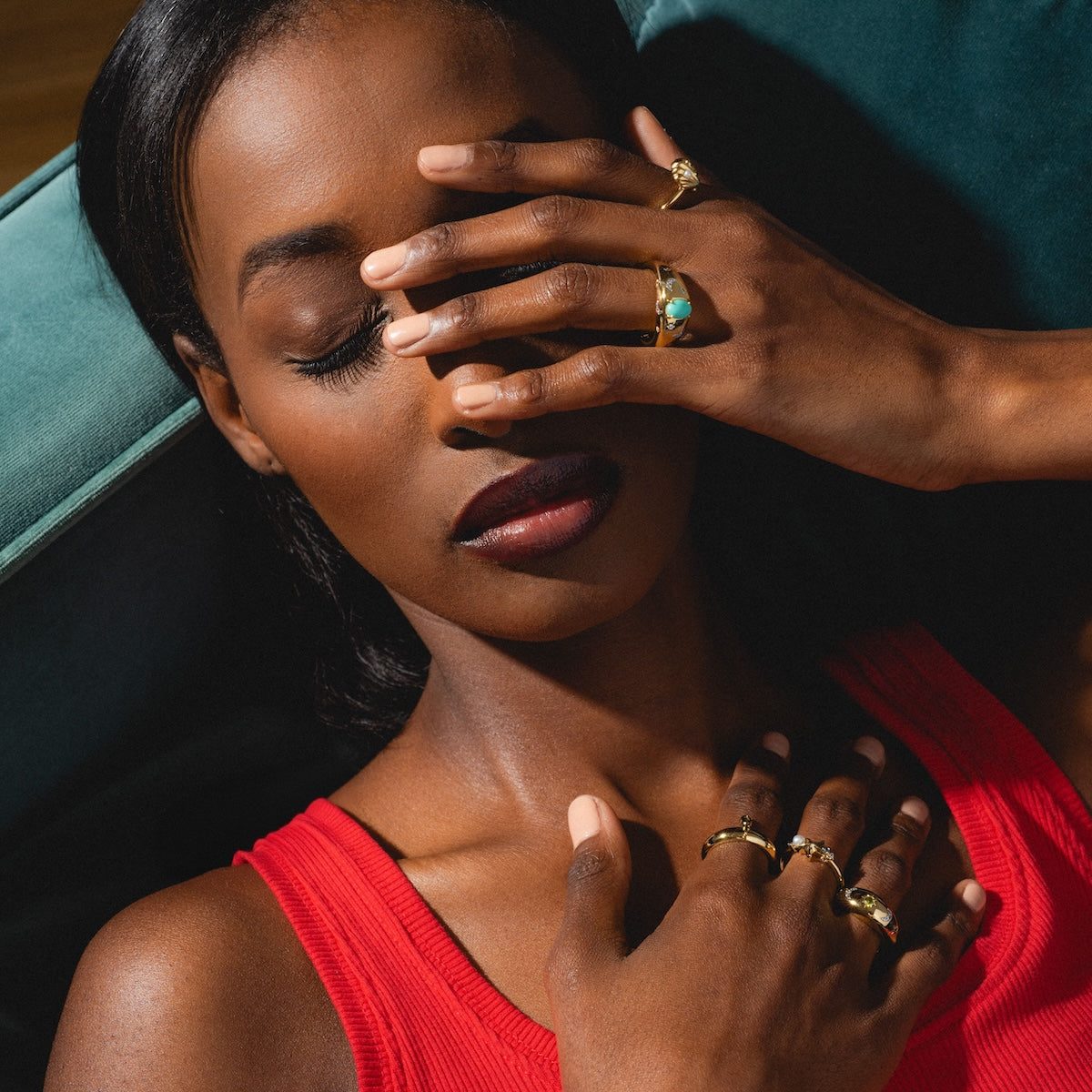 Woman in a red top with multiple gold rings on her fingers, sitting against a dark background.