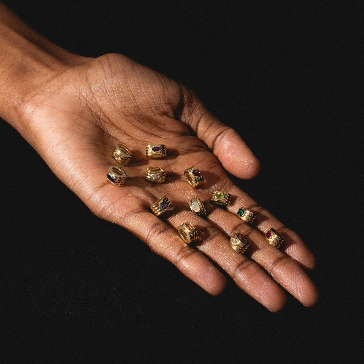 A hand with palm up holds several Solid Gold Bold Birthstone Spacer Charms, each set with colorful gemstones, against a black background.