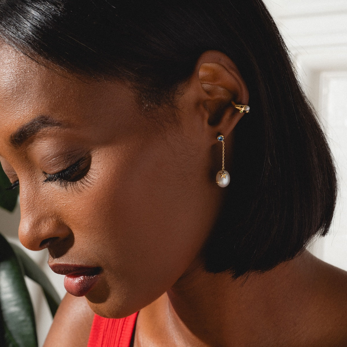 A woman with short, straight black hair wears the Gem Droplet Ear Cuff and a dangling pearl earring. Dressed in a red top, she looks downward, illuminated by soft natural light.