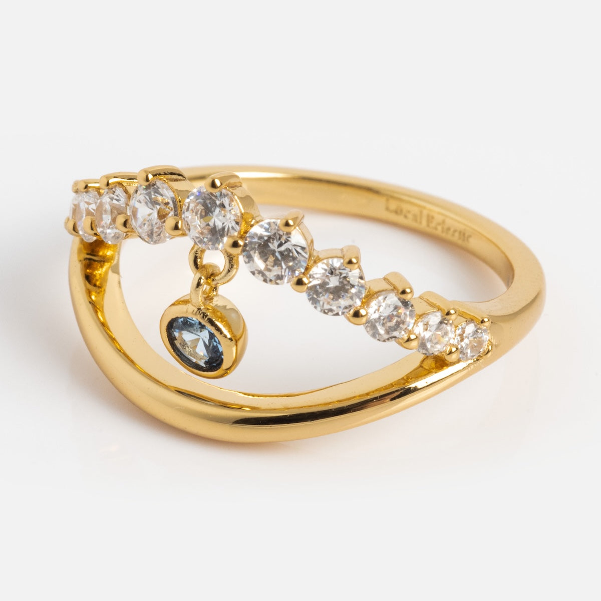 The Mystique Evil Eye Charm Ring is a 14k yellow gold plated ring with a curved band set with seven sparkling gems and a dangling blue gemstone charm at the center.