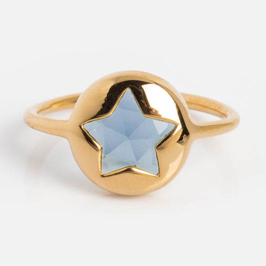 The Maomi Ring is a yellow gold plated ring with a round setting, featuring a faceted light blue star-shaped gemstone at the center, shown on a white background.