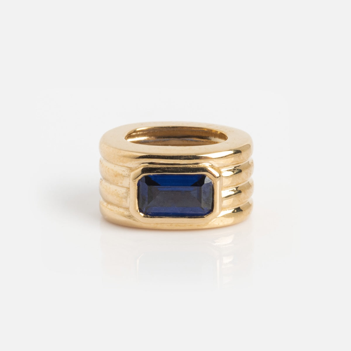 The Solid Gold Bold Birthstone Spacer Charm features a ring-shaped design with three horizontal ridges and a rectangular blue gemstone set in the center, shown against a plain white background.