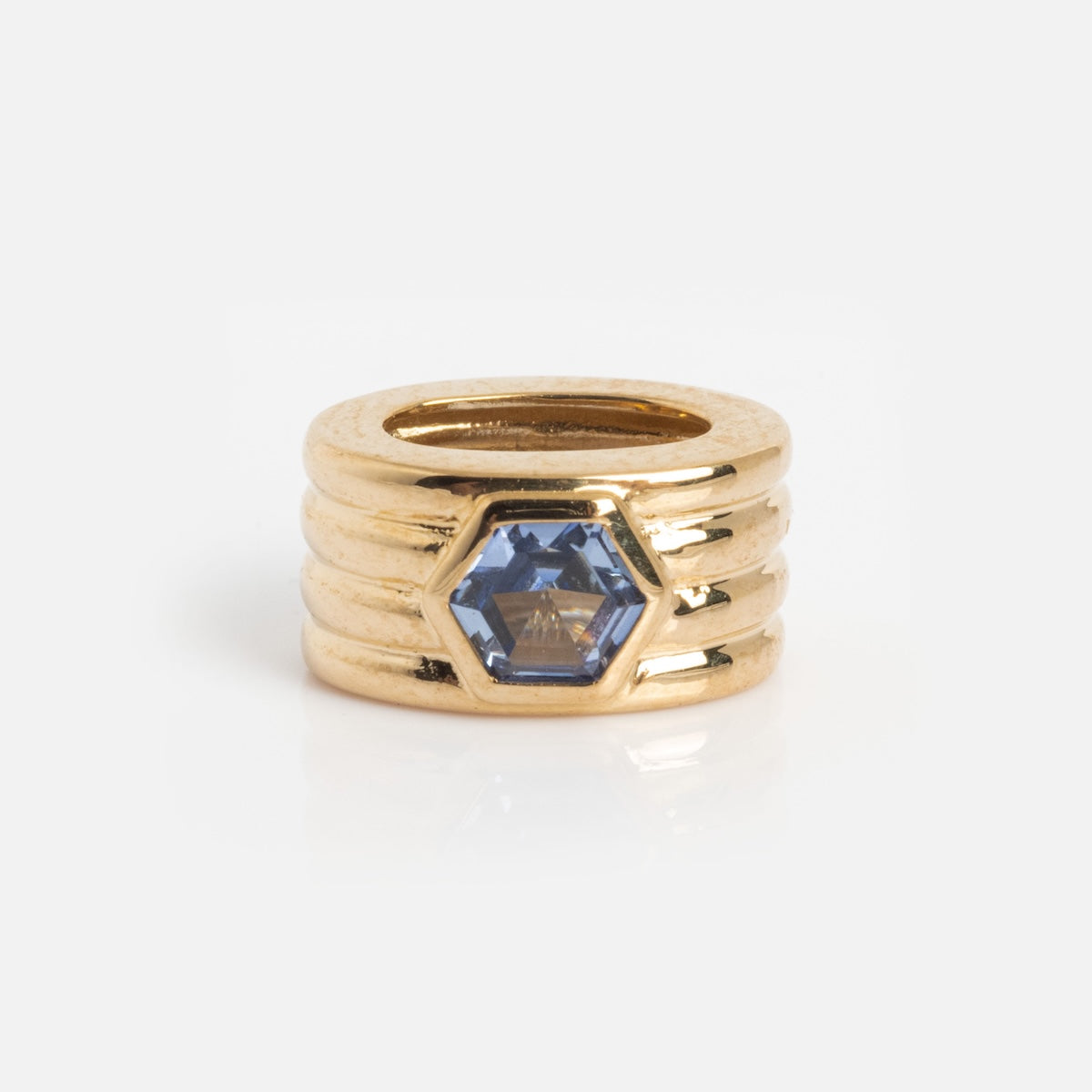 The Solid Gold Bold Birthstone Spacer Charm features a hexagonal blue gemstone set in the center of four stacked, grooved gold bands, displayed against a plain white background.