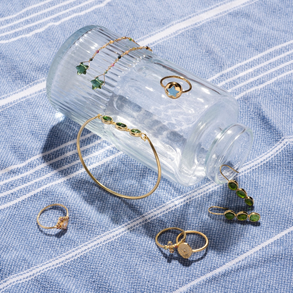 A clear glass jar lies on its side on a blue and white striped cloth, showcasing yellow gold plated jewelry with green and clear gemstones, including the Notre Dame Ring, along with bracelets, earrings, and a necklace.