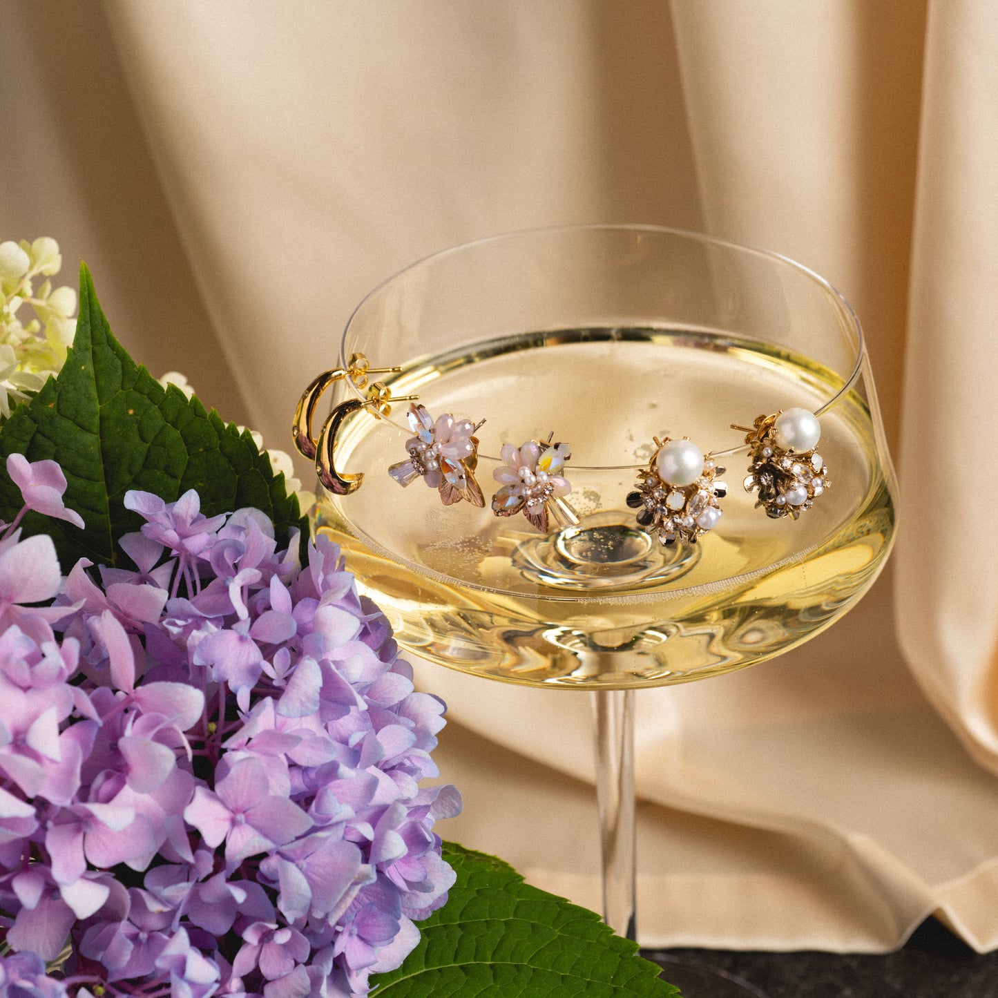 A coupe glass of champagne holds four Luxe Pearl and Crystal Cluster Earrings, each with pearls and gold details, on its rim. Purple hydrangeas and green leaves rest beside the glass on softly draped beige fabric.