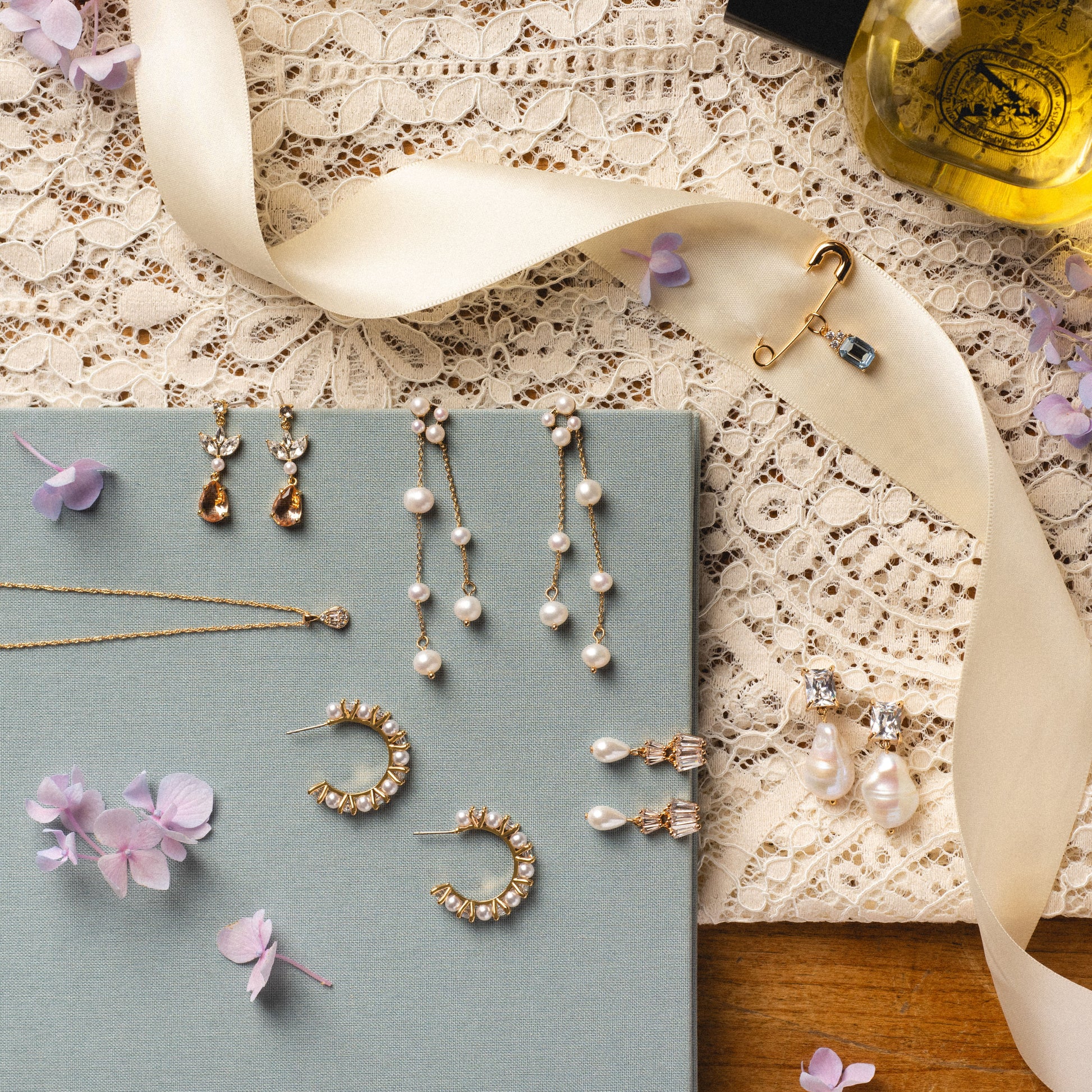 A flat lay of elegant jewelry on lace and fabric, showcasing the Zeta Drop Earrings with freshwater pearls, a gold necklace, brooches, scattered purple flower petals, and a perfume bottle in the corner.