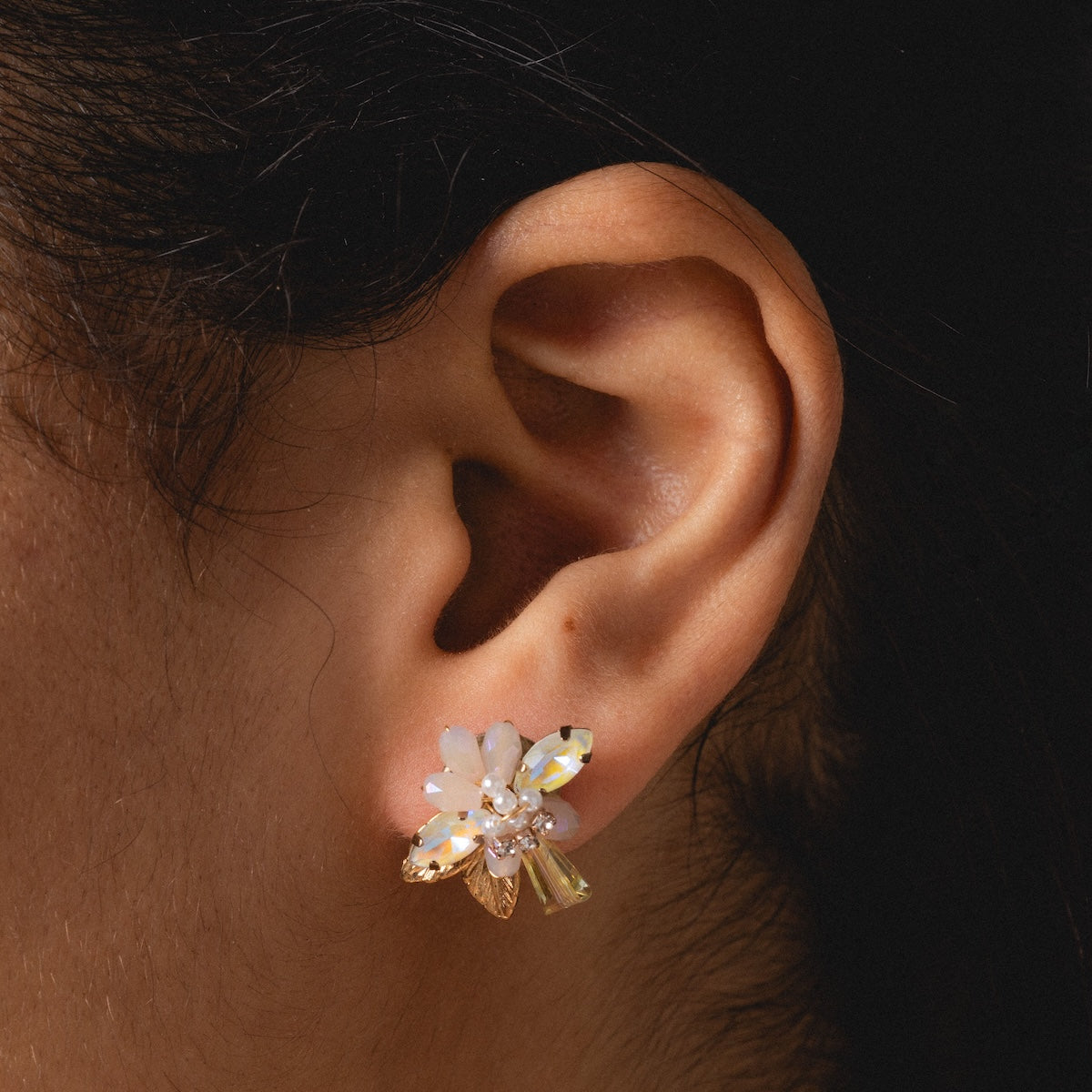 Close-up of an ear wearing Flora Crystal Cluster Earrings, featuring 14k gold plating, white and gold flower petals, and sparkling crystal accents. The dark background is blurred for emphasis.