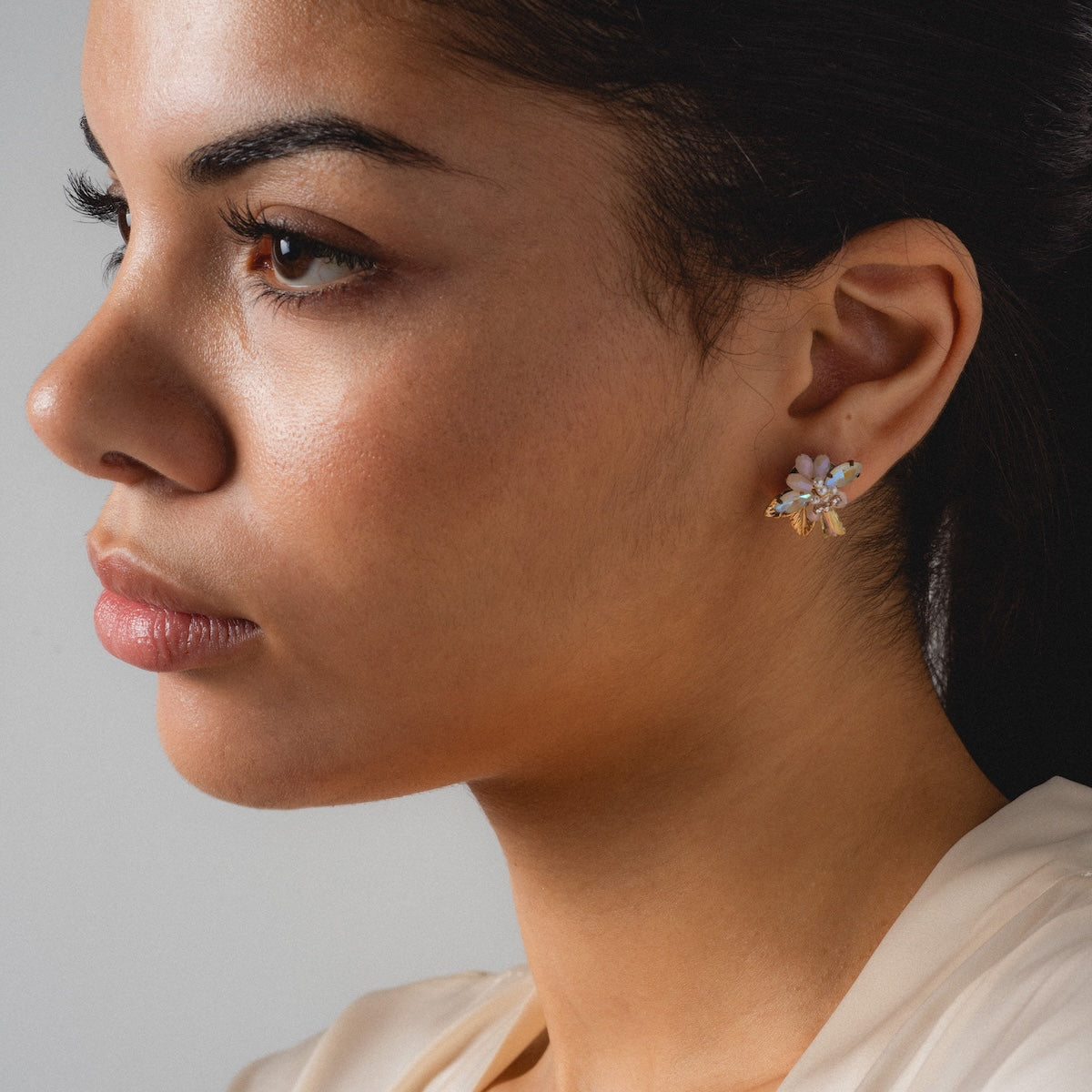 A woman with smooth skin and dark hair looks left, wearing a cream top and Flora Crystal Cluster Earrings. The 14k gold plated crystal accents shimmer against a plain, neutral background.
