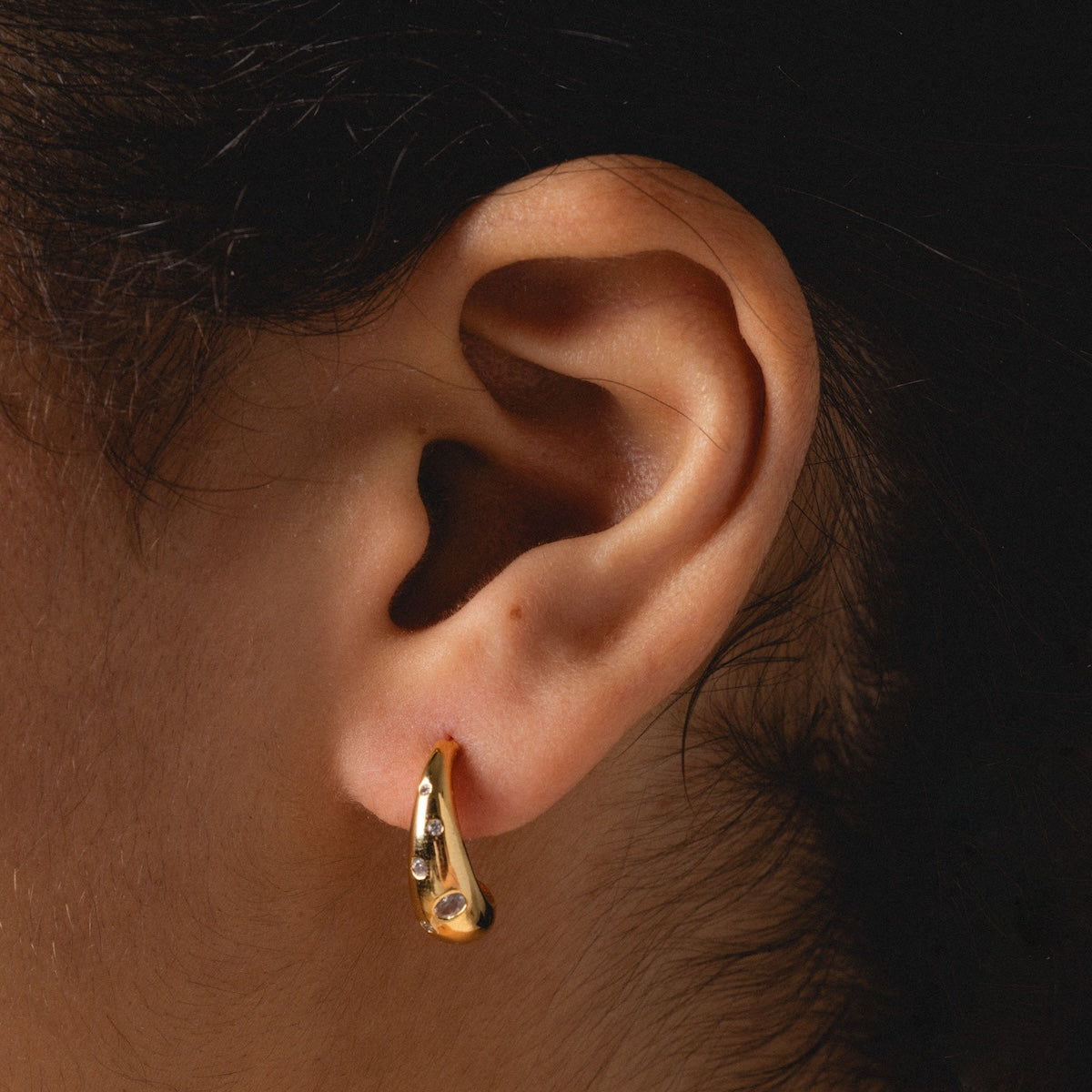 Close-up of a person’s ear wearing the Golden Shimmer Tapered Hoop Earrings. The small gold hoops feature a subtle decorative design, and the dark background highlights their elegant shimmer.
