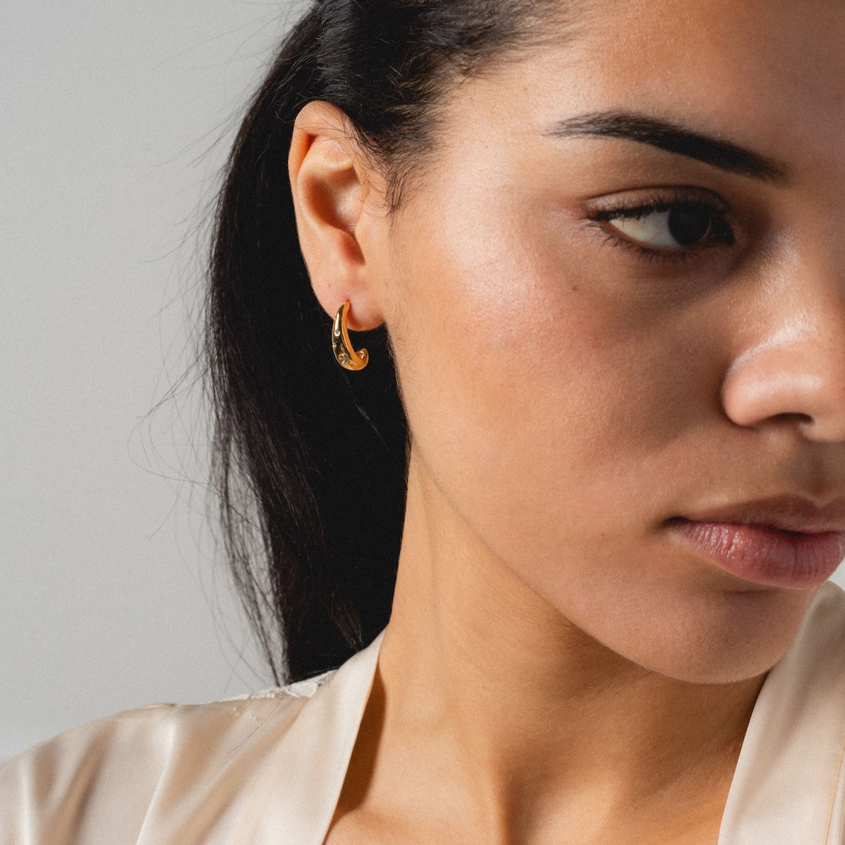 A close-up of a woman with long dark hair wears the Golden Shimmer Tapered Hoop Earrings and a light, silky top, looking slightly downward and to the right against a plain background.