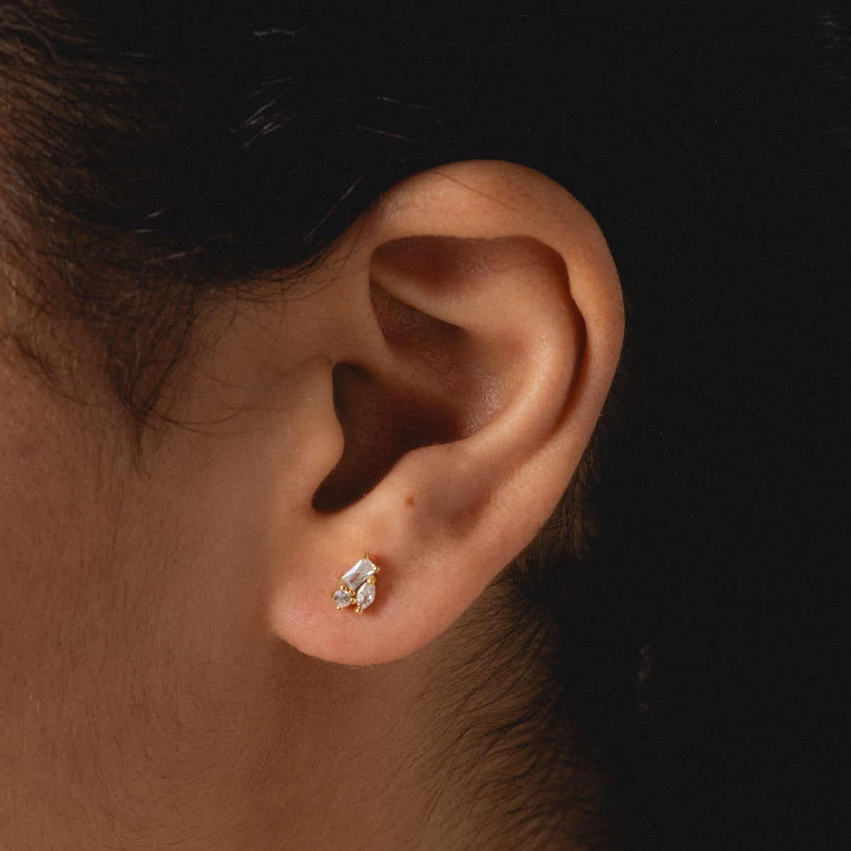 A close-up of a person with short dark hair wearing the Petite Pierre Studs—small, elegant yellow gold plated earrings featuring a rectangular and a round clear gemstone.