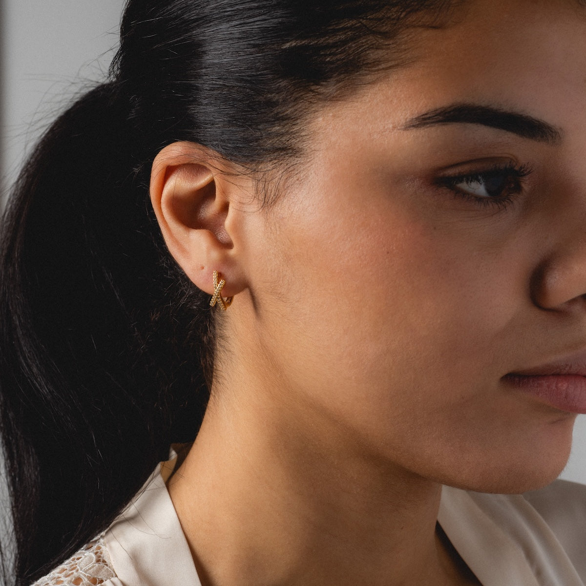 A woman with dark hair in a ponytail wears the Staci Huggies, butterfly-shaped yellow gold earrings. She gazes slightly right, showing only the left side of her face, and is dressed in a cream-colored top.