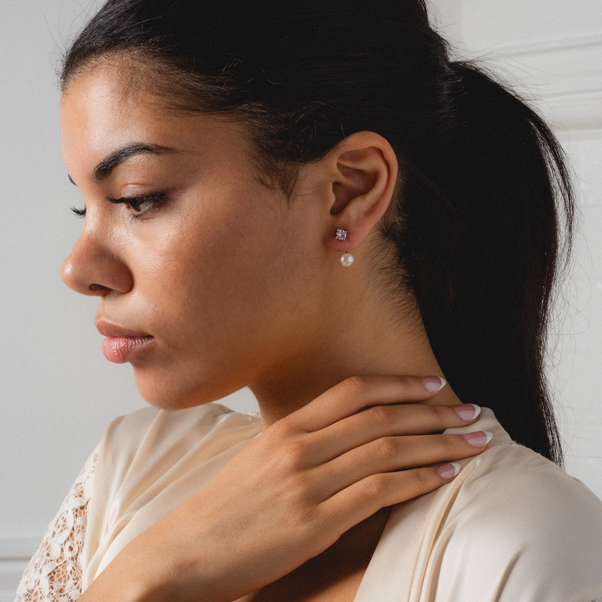 A woman with dark hair in a ponytail wears a cream top and the Aria Earrings, gently touching her neck while gazing downward against a white background.