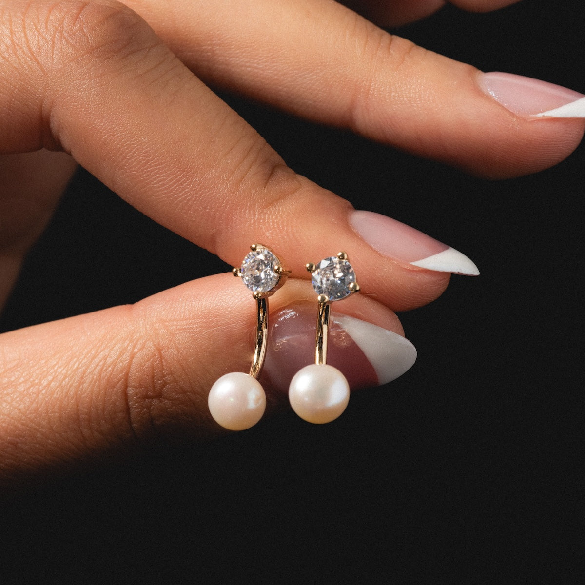 A manicured hand holds the Aria Earrings, showcasing gold-plated accents, a clear gemstone at the top, and a round white freshwater pearl beneath; their elegance stands out against a dark background.