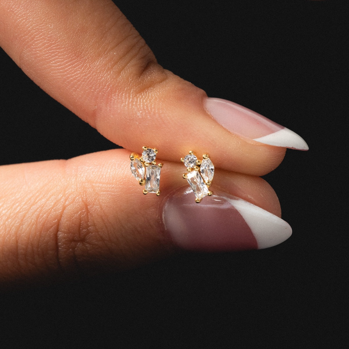 A hand with manicured nails holds the Petite Pierre Studs—yellow gold plated earrings featuring three clear, uniquely shaped CZ gemstones in a cluster design—against a black background.