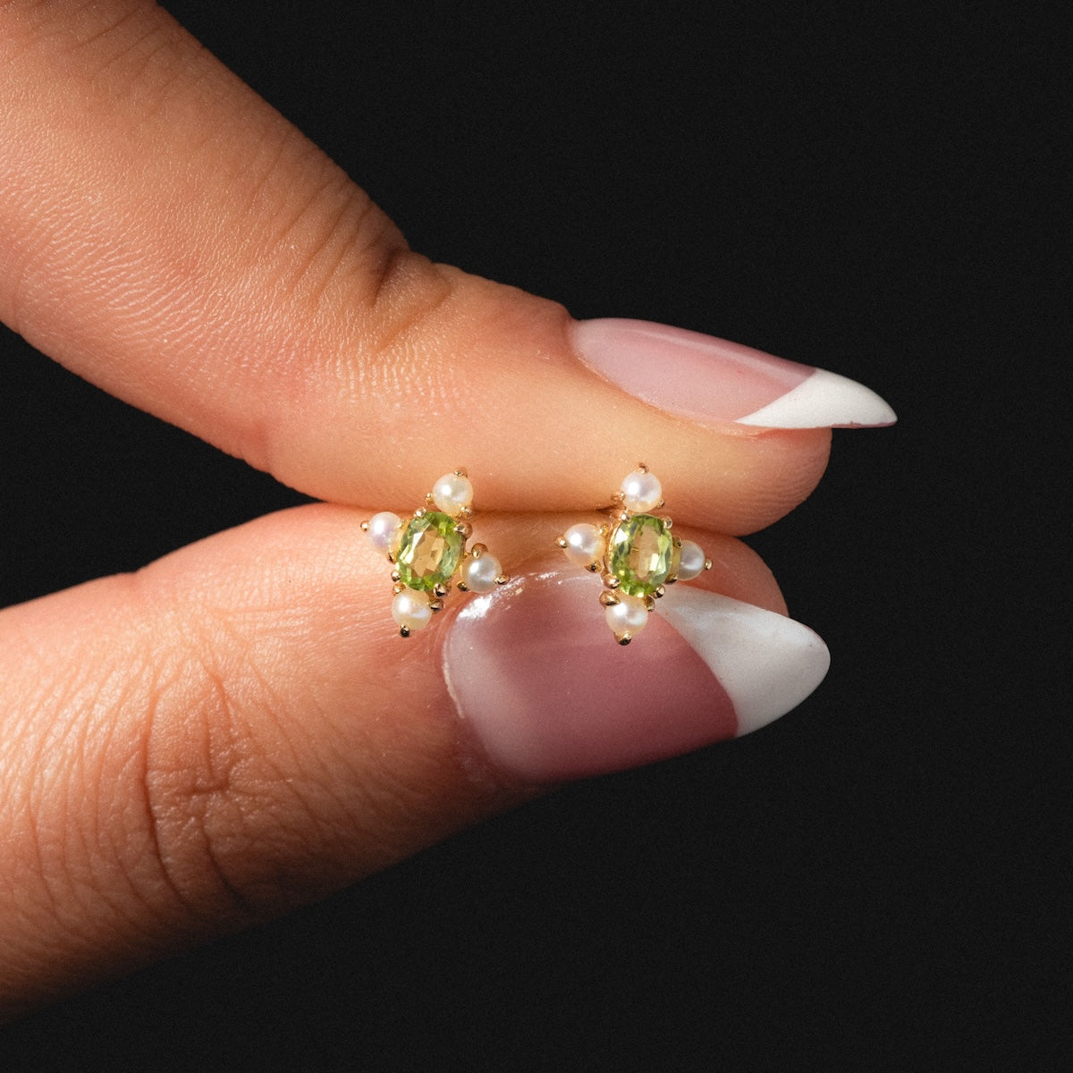A hand with a French manicure displays the Solid Gold 2025 August Capsule Vintage Inspired Peridot Stud Earrings, each featuring an August birthstone and small white pearls, set against a black background.