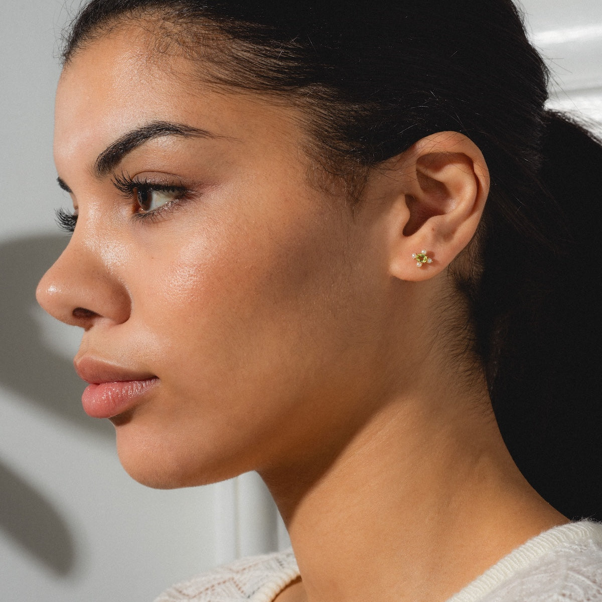 A woman with clear skin and dark hair pulled back in profile against a light background models the Solid Gold 2025 August Capsule Vintage Inspired Peridot Stud Earrings.