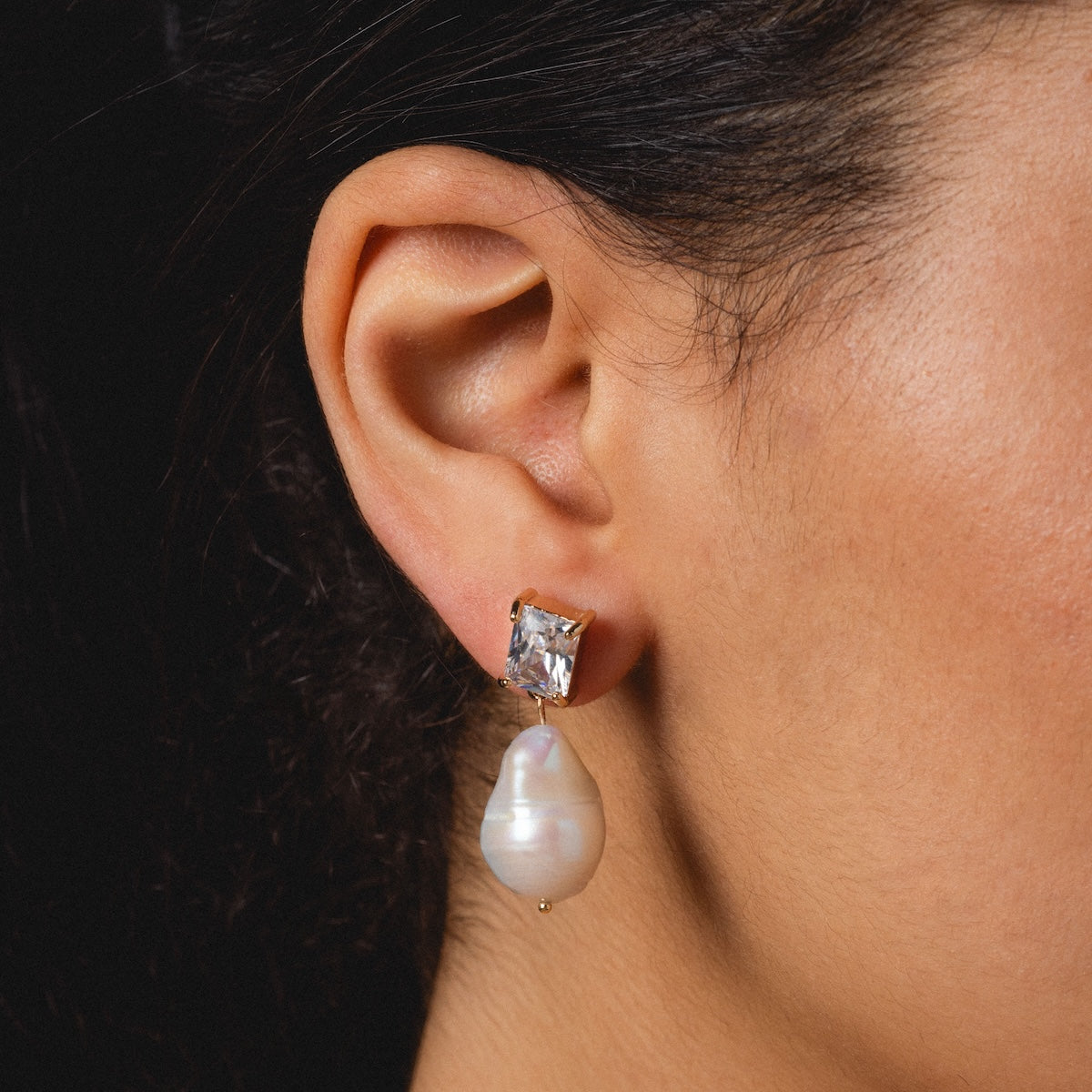 Close-up of a womans ear wearing Zeta Drop Earrings: 14k gold plated with an emerald-cut CZ and a dangling, irregularly-shaped freshwater pearl. Black background.