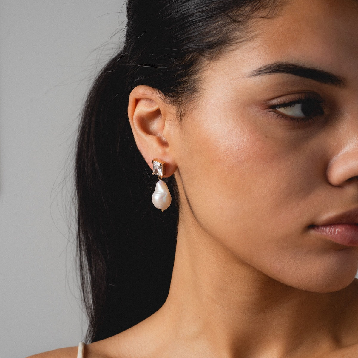 A woman with dark hair wears the Zeta Drop Earrings, featuring freshwater pearls in a silver setting. The image captures the right side of her face and ear against a plain light background.