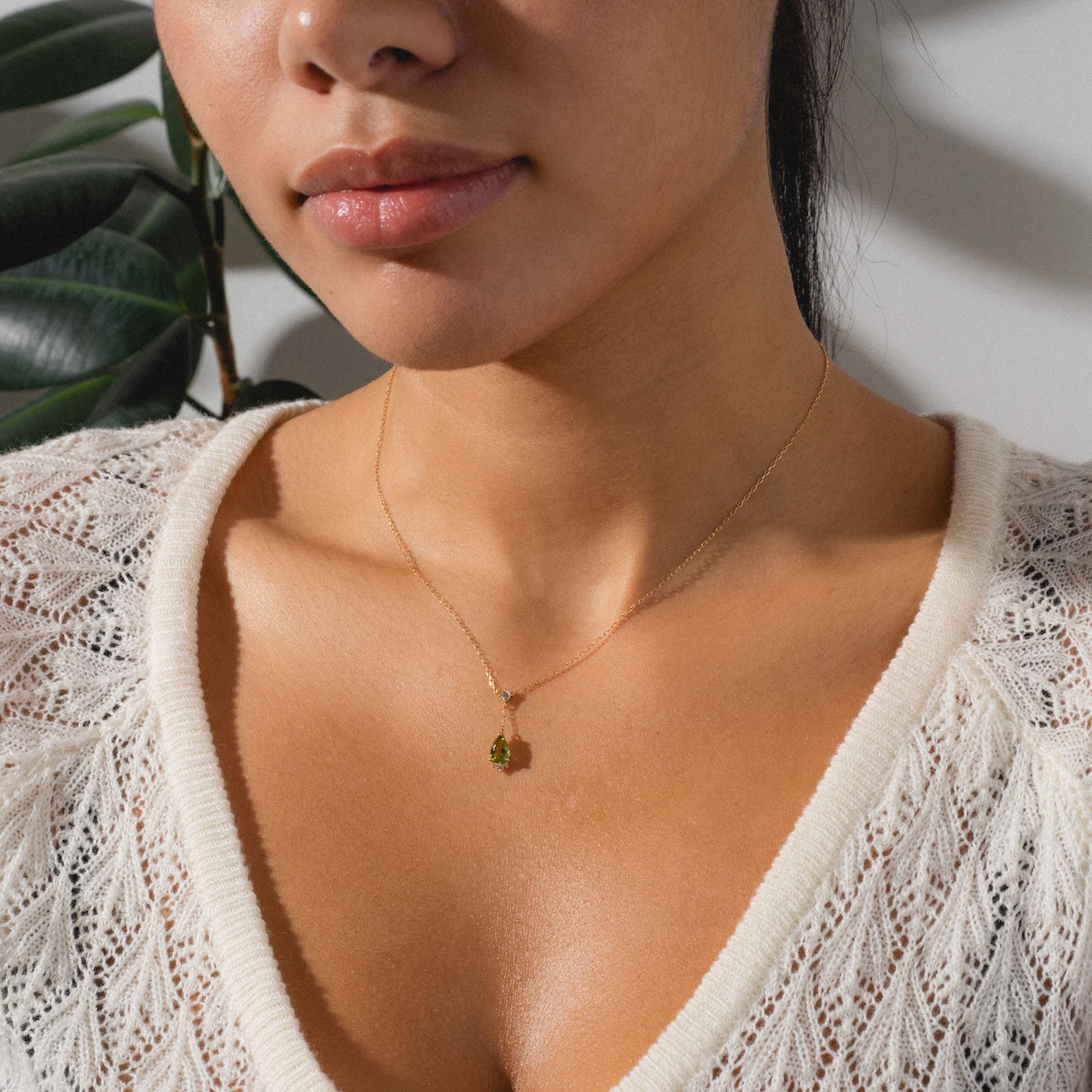 A woman in a white knit top wears the Solid Gold 2025 August Capsule Vintage Inspired Peridot Drop Necklace, featuring an elegant peridot pendant, as she poses in soft natural light beside a leafy plant.