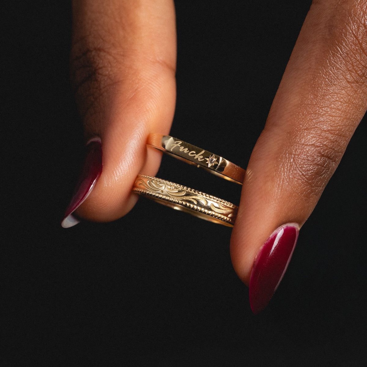 A hand with dark skin and deep red nails holds the Solid Gold Tiny Fuck Filigree Stacking Ring Set: one ring engraved with lucky and a clover, the other adorned with an ornate filigree pattern.
