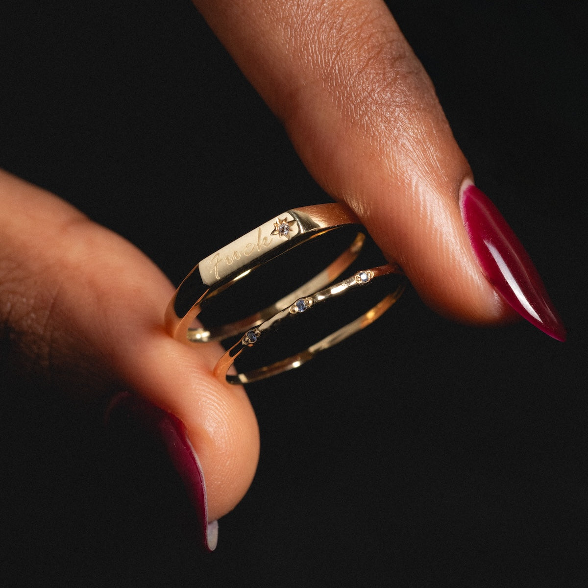 A hand with burgundy nails holds the Solid Gold Tiny Fuck Birthstone Stacking Ring Set—one ring engraved with True and a small diamond, the other accented with tiny diamonds.