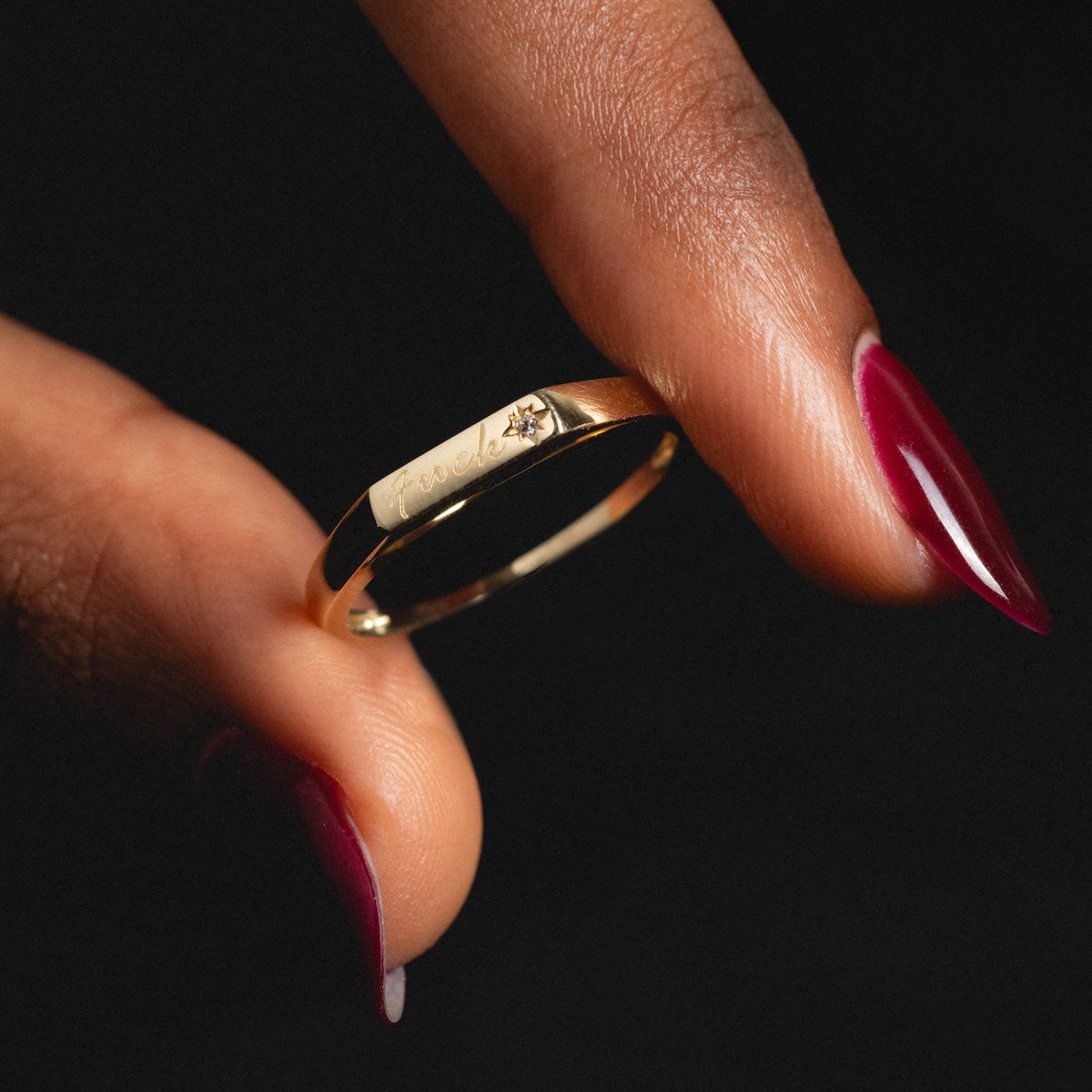 A close-up of a hand with red-painted nails holding the Solid Gold Tiny Fuck Ring, engraved and set with a small diamond, against a dark background.