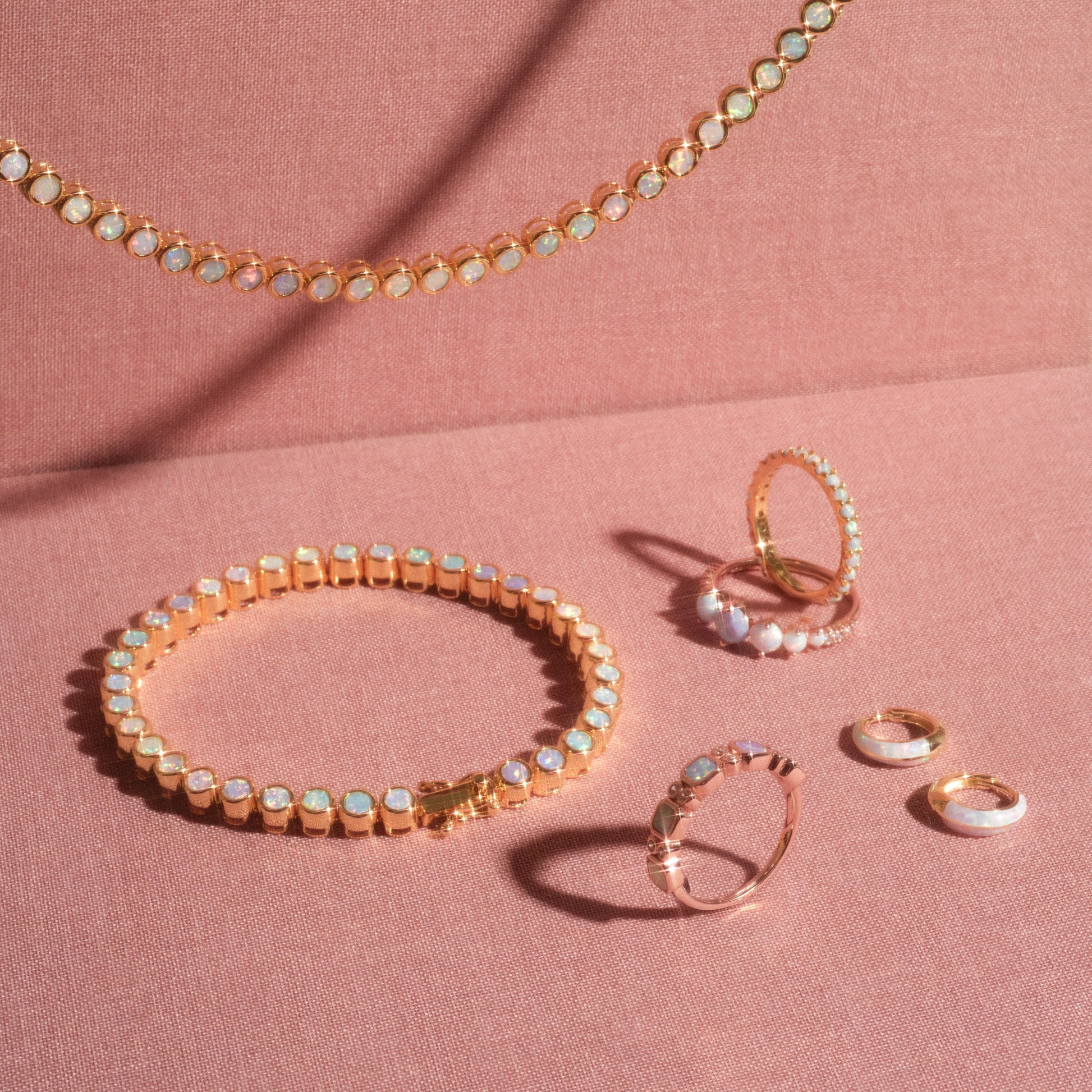 Gold jewelry set with simulated opals—necklace, bracelet, two rings, and three small hoop earrings—displayed on soft pink fabric with warm lighting. Features the 14k rose gold vermeil Rose Gold Ombre Juniper Band.