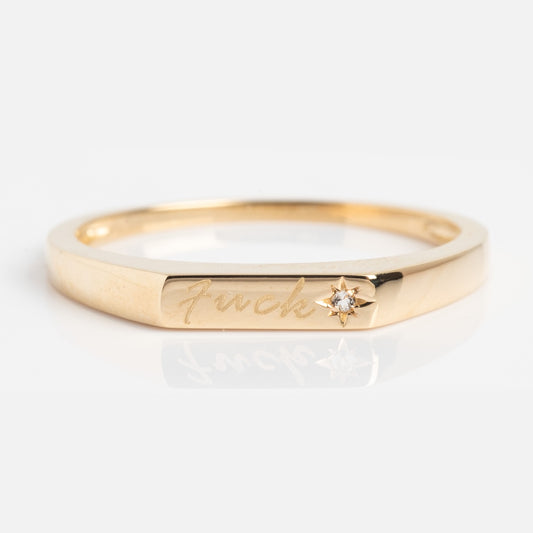 The Solid Gold Tiny Fuck Ring features a slim gold band engraved with “Fuck” in script and accented by a small diamond set in a star detail. Shown here on a white background.