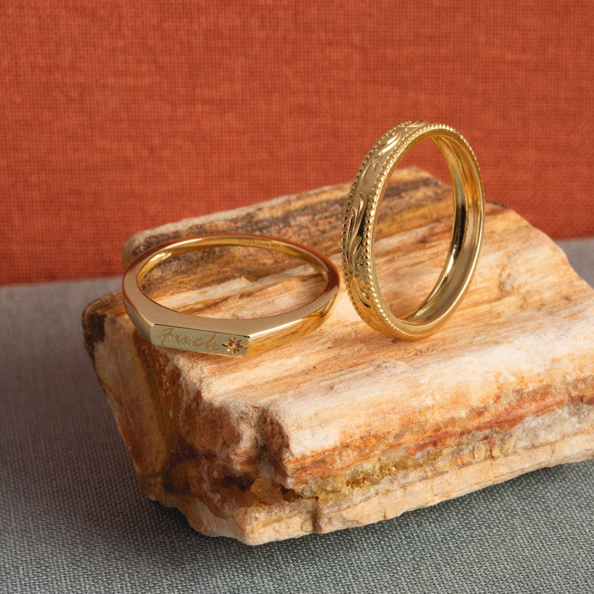 Two solid gold filigree stacking rings sit on textured stone; one features a delicate floral pattern, the other is simpler with a tiny engraved design and “Eternal” inscribed on the band. The background is orange and grey fabric.