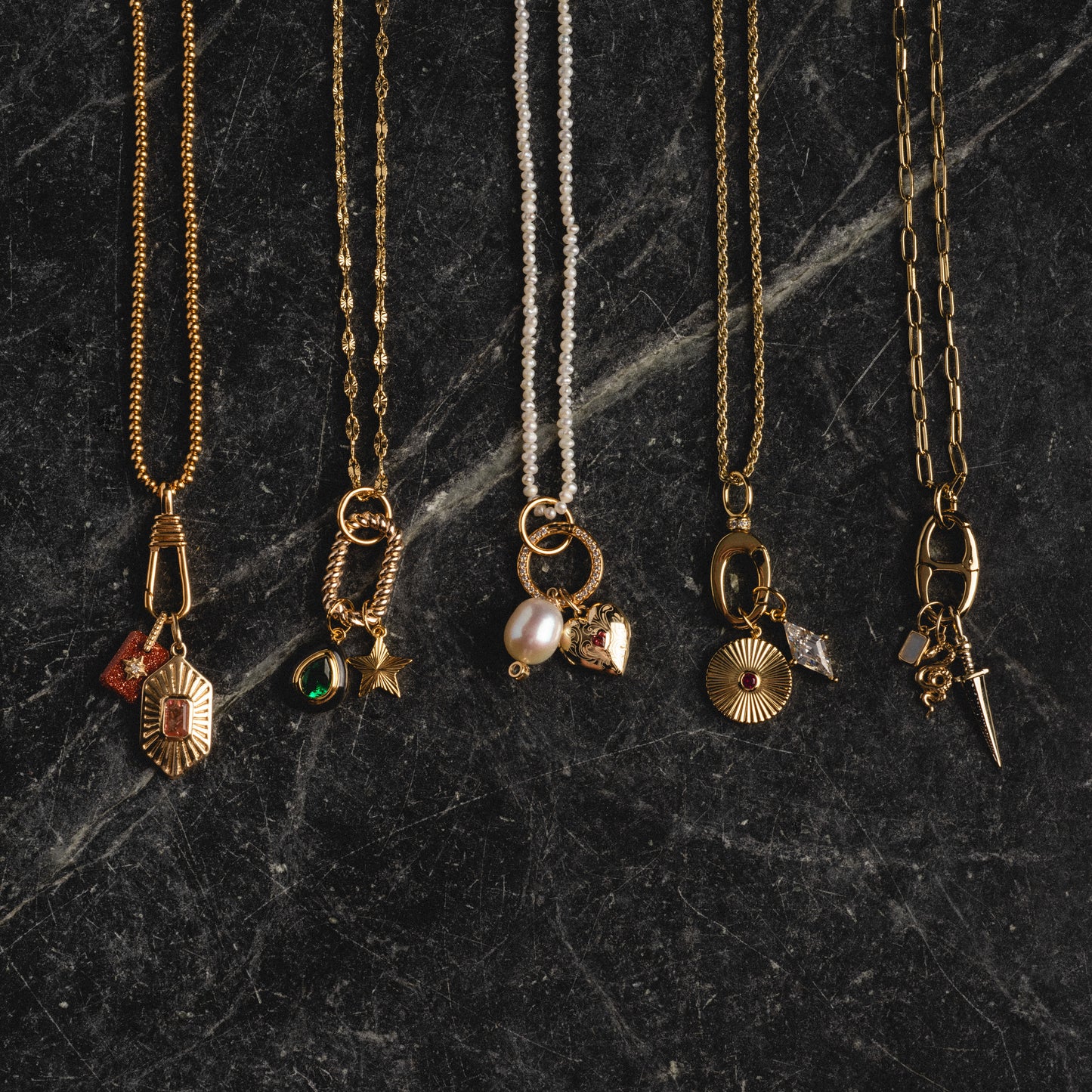 Five Charm Clips with unique pendants, including gold vermeil, pearl, and a small dagger, are displayed side by side on a dark, textured stone surface.