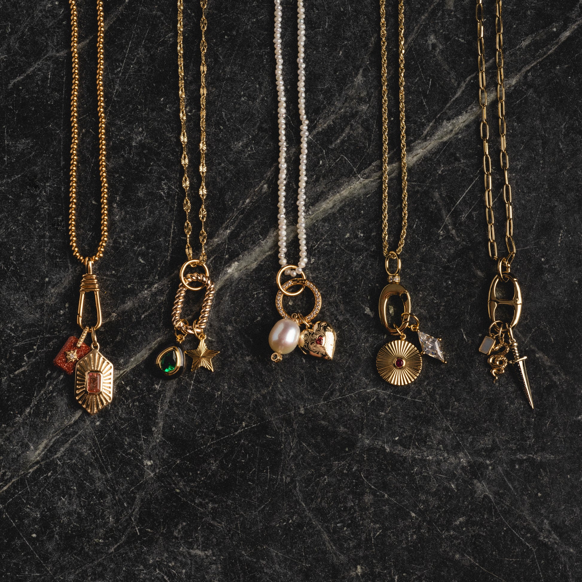 Five Charm Clips with unique pendants, including gold vermeil, pearl, and a small dagger, are displayed side by side on a dark, textured stone surface.