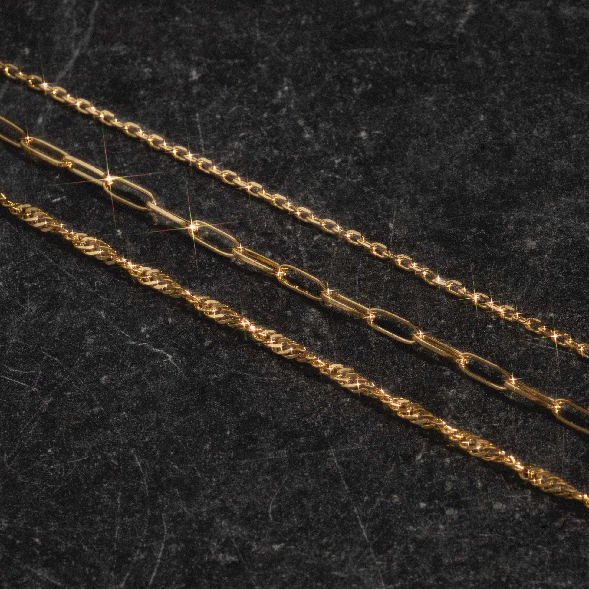 The Oval Link Chain, a gold-plated necklace, is arranged diagonally on a dark textured surface, with light reflecting off the metal for sparkling highlights—a true charm collection.