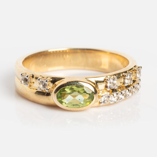 The Solid Gold Peridot Accent Band (Sample Size 7) features an oval green peridot center stone, accented by small clear stones along the band, and is displayed against a white background.