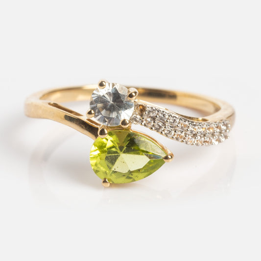 The Solid Gold Peridot Toi et Moi Wrap Ring, sample size 7, features a pear-shaped green peridot, a round clear stone, and a curved gold band with small clear stones on a white background.
