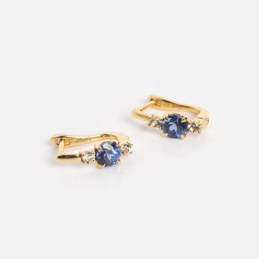 Solid Gold Tanzanite and White Topaz Huggie Hoops, featuring blue center stones and small clear side stones, displayed on a white background.