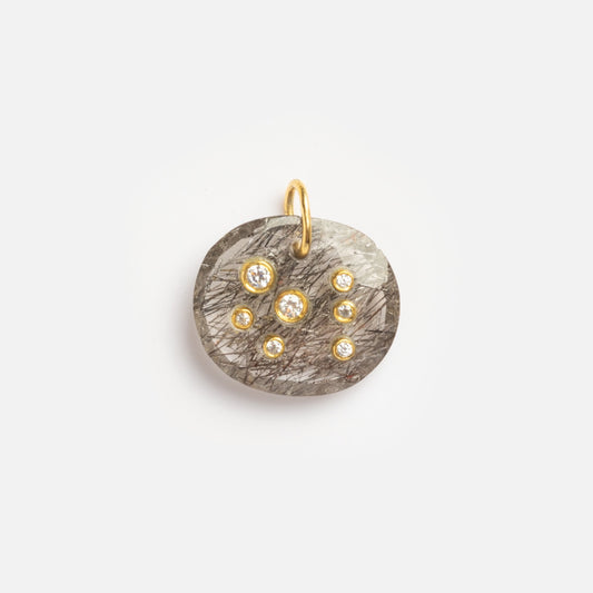 The Black Rutile Scorpio Constellation Charm Sample is a round gray stone pendant with black streaks, seven small clear gemstones arranged in a constellation pattern, and a gold loop for attachment.