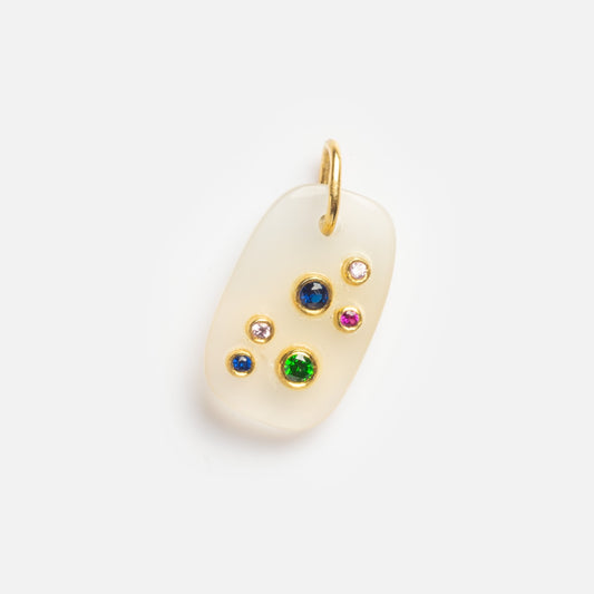 The White Chalcedony Taurus Constellation Charm Sample is a rectangular white pendant with rounded corners, a gold loop for hanging, and six small blue, green, pink, and clear gemstones arranged asymmetrically on its surface.