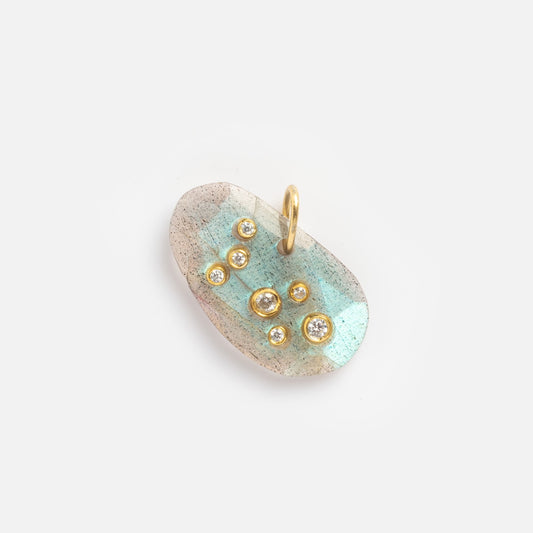 The Labradorite Virgo Constellation Charm Sample is an oval pendant with a light blue-green iridescent surface, adorned with seven small round diamonds in gold bezels and a gold bail on top against a plain white background.