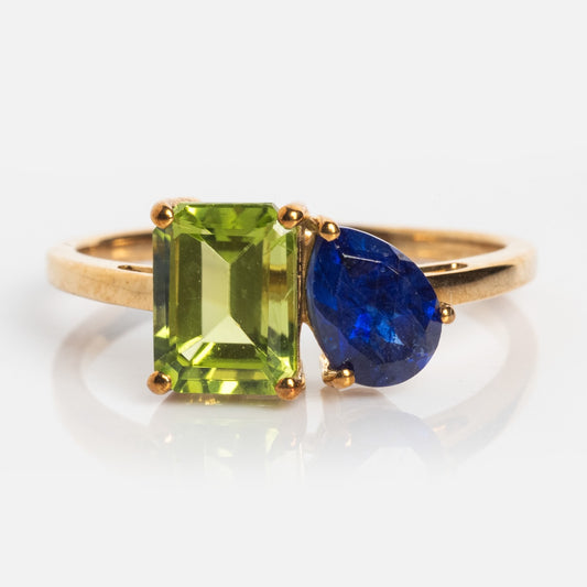 The Customizable Toi et Moi Ring Peridot and Sapphire (Sample Size 8) features a gold band with a rectangular green peridot and a teardrop blue sapphire set side by side, shown on a white background.