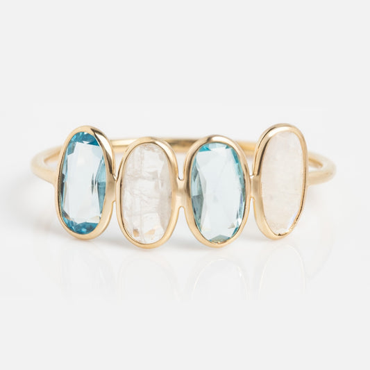 The Solid Gold Daybreak Moonstone and Blue Topaz Ring features four vertically-set oval stones—alternating light blue topaz and translucent white moonstones—crafted in 9k yellow gold, shown on a white background.