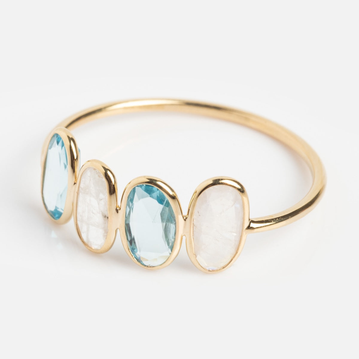 The Solid Gold Daybreak Moonstone and Blue Topaz Ring features four oval gemstones—alternating light blue topaz and translucent white moonstone—in bezel settings on a simple 9k yellow gold band, shown against a plain white background.
