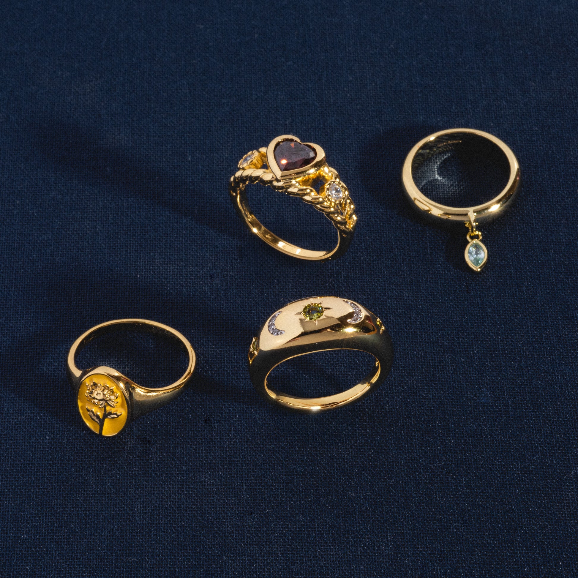 Four 14k gold plated rings on dark blue fabric: the Bold Heart Statement Ring with a heart-shaped stone and diamonds, a ring with a dangling teardrop gem, one with a raised tree design, and one featuring a small green stone in the center.